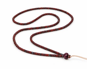May include: A long necklace composed of small, cylindrical dark red beads with occasional metallic grey accents. The strand features a single, larger, smooth dark red bead near the end, which is finished with a light tan cord extension.