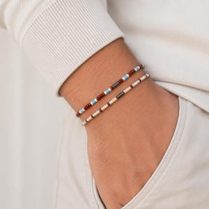 Könnte beinhalten: Zwei zarte Perlenarmbänder am Handgelenk. Das obere Armband besteht aus dunkelroten, hellblauen und weißen Perlen mit einer zentralen dunkelgrauen zylindrischen Perle. Das untere Armband besteht aus cremefarbenen, hellblauen und dunkelroten Perlen.