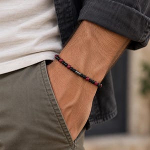 May include: A beaded bracelet featuring a repeating pattern of small, round black and deep red beads. The design is accented by a single, slender, cylindrical dark metallic bead positioned centrally among the smaller spherical beads.