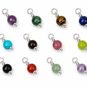 Op de afbeelding: Een collectie van twaalf individuele sieradenbedels, elk voorzien van een enkele ronde edelsteenkraal in diverse kleuren zoals paars, groen, bruin, blauw, turkoois, zwart, rood en lichtgroen, geaccentueerd met zilverkleurige metalen onderdelen.