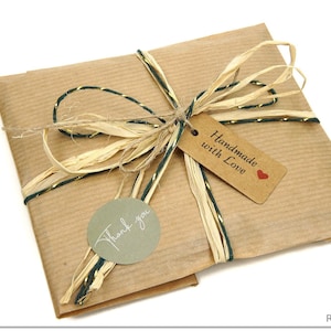 K&ouml;nnte beinhalten: Ein quadratisches Paket in braunem Kraftpapier, verziert mit einer Schleife aus nat&uuml;rlichem Bast und dunkelgr&uuml;ner Schnur. Ein rechteckiges braunes Etikett tr&auml;gt die Aufschrift Handmade with Love mit einem roten Herz und ein runder salbeigr&uuml;ner Aufkleber sagt Thank you.