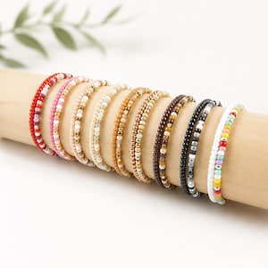 Könnte beinhalten: Eine Sammlung von elf dünnen Perlenarmbändern, die in einer Reihe angeordnet sind. Die Armbänder bestehen aus kleinen, runden Glasperlen in verschiedenen Farben, darunter Rot, Rosa, Beige, Hellbraun, Braun, Schwarz, Grau und ein mehrfarbiges Band.