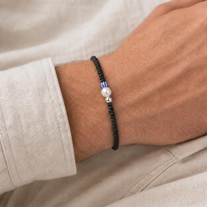 Könnte beinhalten: Ein Perlenarmband mit kleinen mattschwarzen Perlen, einer blau-weiß gestreiften Perle, einer weißen perlmuttartigen Perle und einer kleinen silberfarbenen Metallperle. Das Armband ist auf einer flexiblen Schnur aufgezogen und liegt auf beigem Stoff.