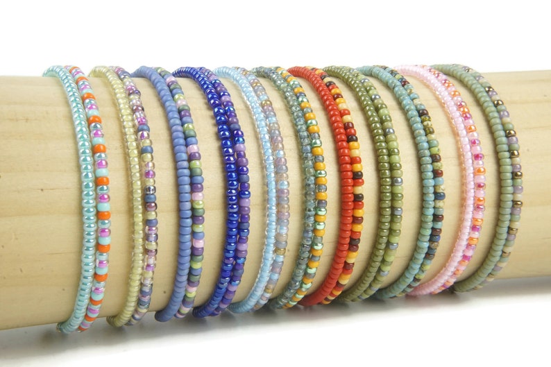 K&ouml;nnte beinhalten: Eine Sammlung d&uuml;nner, elastischer Armb&auml;nder aus kleinen, runden Glasperlen in verschiedenen Farben wie Hellblau, Orange, Violett, Gelb, Rot, Gr&uuml;n und Rosa, die in einer Reihe auf einem zylindrischen Holzst&auml;nder pr&auml;sentiert werden.