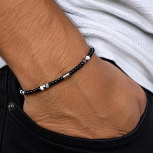 May include: A beaded bracelet featuring a repeating pattern of small black spherical beads, interspersed with white beads and metallic silver-toned cylindrical accents, including a central elongated silver-toned metal tube bead.