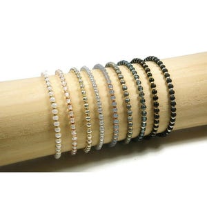 Peut inclure: Une collection de bracelets de perles de différentes couleurs, dont blanc, rose, or, argent, gris et noir. Les bracelets sont présentés sur une cheville en bois, mettant en valeur leur design délicat et leurs palettes de couleurs variées. Chaque bracelet est fait de petites perles.