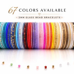Op de afbeelding: Een verzameling 2 mm glazen kralenarmbanden in een regenboog aan kleuren. De afbeelding toont een verscheidenheid aan armbanden in tinten groen, blauw, paars, roze, oranje, geel en meer. De tekst luidt "67 KLEUREN BESCHIKBAAR" en "2MM GLAS KRAAL ARMBANDEN."