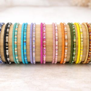 Könnte beinhalten: Eine Sammlung von Perlenarmbändern in verschiedenen Farben, darunter Blau, Schwarz, Türkis, Lila, Rosa, Orange, Grün, Gelb, Gold und Silber. Die Armbänder werden auf einem Holzdübel präsentiert und zeigen ihre vielfältigen Farbtöne und Designs.