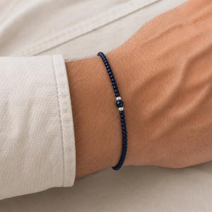 Op de afbeelding: Een dunne armband bestaande uit kleine, ronde, donkerblauwe kralen. Het ontwerp heeft een enkele, iets grotere, gefacetteerde donkerblauwe kraal in het midden, geflankeerd door twee kleine, gepolijste zilverkleurige metalen tussenstukjes aan weerszijden.