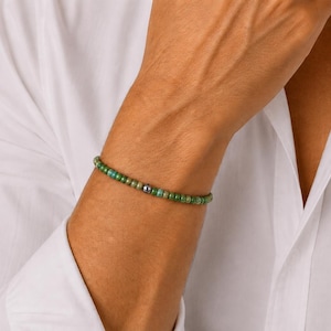 May include: A beaded bracelet featuring small, round, green and turquoise beads. A single, darker bead is centered on the bracelet. The bracelet is worn on a person's arm, against the backdrop of a white shirt.