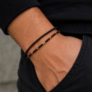 Könnte beinhalten: Zwei dünne Perlenarmbänder, die zusammen getragen werden. Ein Armband besteht aus kleinen, gleichmäßigen schwarzen Perlen. Das zweite Armband zeigt schwarze Perlen, die mit kleinen orangefarbenen Akzenten und einer silberfarbenen Metallperle versehen sind.