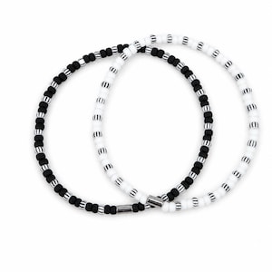 Könnte beinhalten: Zwei kreisförmige Armbänder aus kleinen Perlen. Ein Armband besteht aus einfarbigen schwarzen Perlen im Wechsel mit schwarz-weiß gestreiften Perlen. Das zweite Armband besteht aus weißen Perlen im Wechsel mit schwarz-weiß gestreiften Perlen.