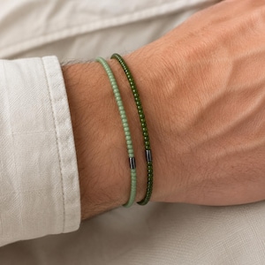 Könnte beinhalten: Zwei dünne Perlenarmbänder, die zusammen getragen werden. Ein Armband besteht aus kleinen, hellen salbeigrünen Perlen, das andere aus dunkelgrünen Waldgrün-Perlen. Jedes Armband enthält eine einzelne, kleine, zylindrische Metallperle in Dunkelgrau.