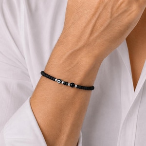 May include: A black beaded bracelet with silver details and an evil eye charm. The bracelet is worn on a wrist, against a white shirt. The bracelet has small, round black beads and a central evil eye bead.
