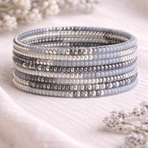 May include: A collection of beaded bracelets in grey and silver tones. The bracelets are composed of small, round beads and are arranged in a spiral pattern. The bracelets are presented on a white surface.