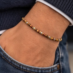 May include: A beaded bracelet with a mix of brown, gold, and silver beads. The bracelet is worn on a wrist, with a dark blue sweater and denim jeans in the background. The bracelet is a simple, elegant accessory.