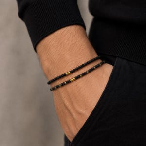 Könnte beinhalten: Zwei dünne Perlenarmbänder mit kleinen mattschwarzen, kugelförmigen Perlen. Jedes Armband ist mit einer einzelnen glatten, zylindrischen, goldfarbenen Metallperle verziert. Ein Armband besteht nur aus schwarzen Perlen, das andere enthält metallische Akzente.