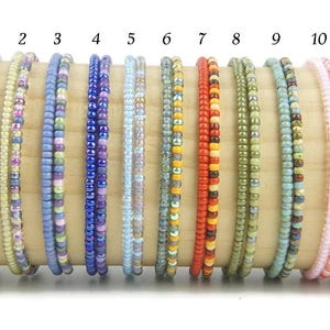 K&ouml;nnte beinhalten: Elf d&uuml;nne Armb&auml;nder aus kleinen, bunten Glasperlen, nummeriert von eins bis elf. Jedes Armband weist eine einzigartige Farbkombination auf, darunter Blau-, Orange-, Gelb-, Rot-, Gr&uuml;n- und Rosat&ouml;ne, die in einer Reihe angeordnet sind.