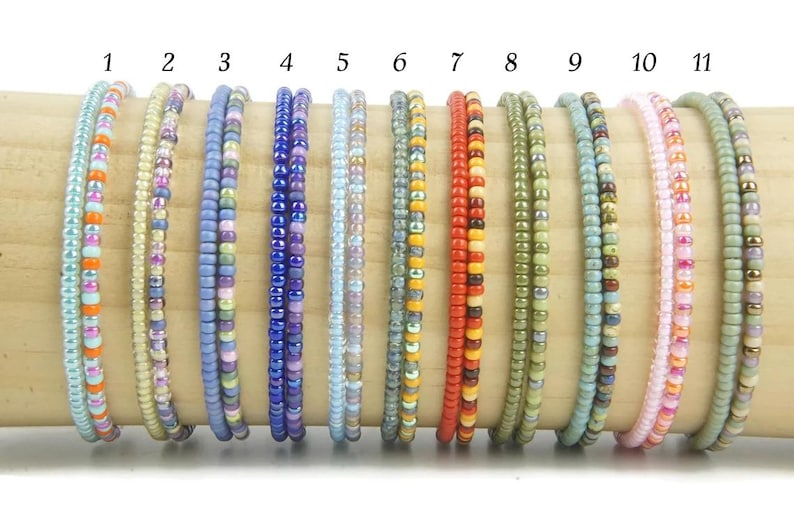 K&ouml;nnte beinhalten: Elf d&uuml;nne Armb&auml;nder aus kleinen, bunten Glasperlen, nummeriert von eins bis elf. Jedes Armband weist eine einzigartige Farbkombination auf, darunter Blau-, Orange-, Gelb-, Rot-, Gr&uuml;n- und Rosat&ouml;ne, die in einer Reihe angeordnet sind.