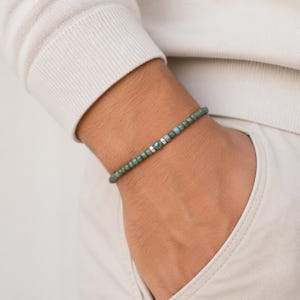 Könnte beinhalten: Ein Perlenarmband mit kleinen, runden, salbeigrünen Steinen, die in einer durchgehenden Linie angeordnet sind. Das Design wird durch zwei kleine, polierte, silberfarbene zylindrische Abstandshalter in der Mitte akzentuiert.