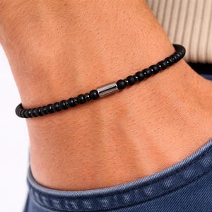 Peut inclure: Bracelet de perles noires avec un accent cylindrique argenté. Le bracelet est composé de petites perles rondes noires mates. Le bracelet est porté au poignet.