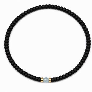 Op de afbeelding: Zwart kralenarmband met een lichtblauwe edelsteen en goudkleurige accenten. De armband is gemaakt van kleine, ronde zwarte kralen, met een enkele lichtblauwe edelsteen in het midden. De armband heeft een omtrek van ongeveer 18 cm.