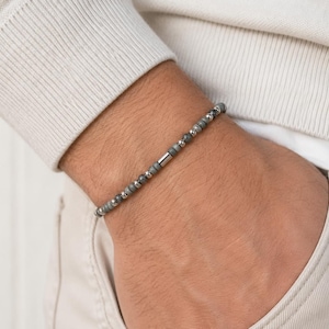 Könnte beinhalten: Ein Perlenarmband mit abwechselnden kleinen grauen Steinperlen und polierten silberfarbenen Metallperlen. Das Design enthält ein zentrales zylindrisches silberfarbenes Metallelement, das ein gleichmäßiges und wiederkehrendes Muster bildet.