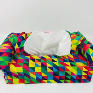 May include: A fabric tissue box cover with a vibrant geometric pattern in shades of red, yellow, green, and blue. The cover is shaped like a small sofa, with rounded armrests and a white tissue emerging from the top.
