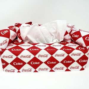 May include: A red and white tissue box cover shaped like a couch with pillows. The cover is decorated with a repeating pattern of red diamonds with "Coke" and "Coca-Cola" printed in white.