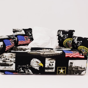 May include: A black tissue box cover with a camouflage pattern featuring American flags, eagles, tanks, and military insignia. The cover has four pillow-like flaps that are also decorated with the same camouflage pattern.