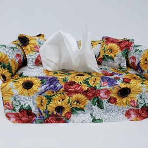 May include: A tissue box cover with a floral design featuring sunflowers, roses, and lavender. The cover is made of fabric and has a white background with a gray pattern. The cover has four pillows with a sunflower and rose design.