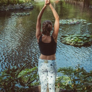 May include: A woman in a black tank top and white patterned leggings is practicing yoga by a pond. She has her arms raised above her head with her hands together in a prayer position.