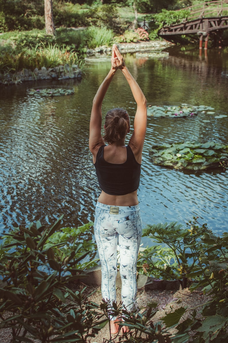 May include: A woman in a black tank top and white patterned leggings is practicing yoga by a pond. She has her arms raised above her head with her hands together in a prayer position.