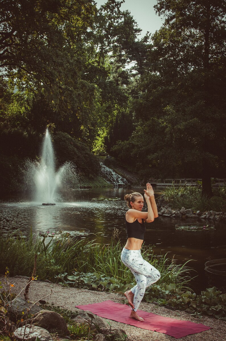 May include: A woman in a black sports bra and white patterned leggings performs a yoga pose on a red mat outdoors. She is standing on one leg with her other leg bent and her arms raised above her head. The background features a pond with a fountain and lush greenery.