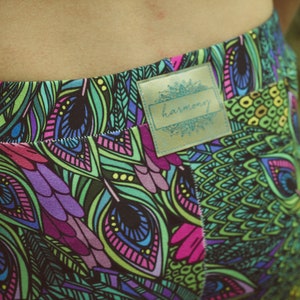 May include: Close-up of colorful leggings with a vibrant peacock feather pattern in shades of green, purple, pink, and blue. The leggings have a label that says "harmony".