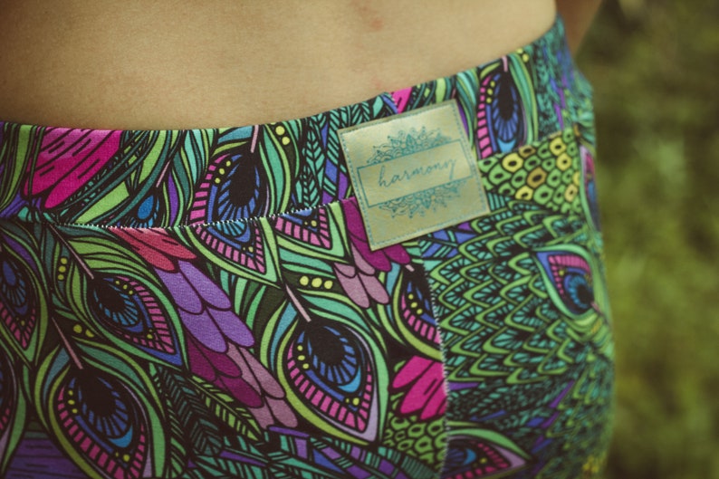 May include: Close-up of colorful leggings with a vibrant peacock feather pattern in shades of green, purple, pink, and blue. The leggings have a label that says "harmony".