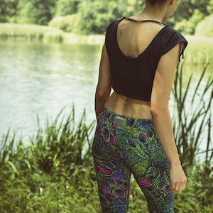 May include: A person wearing a black crop top and patterned leggings. The leggings feature a vibrant peacock feather design in shades of green, purple, and pink. The background shows a lake and lush greenery.