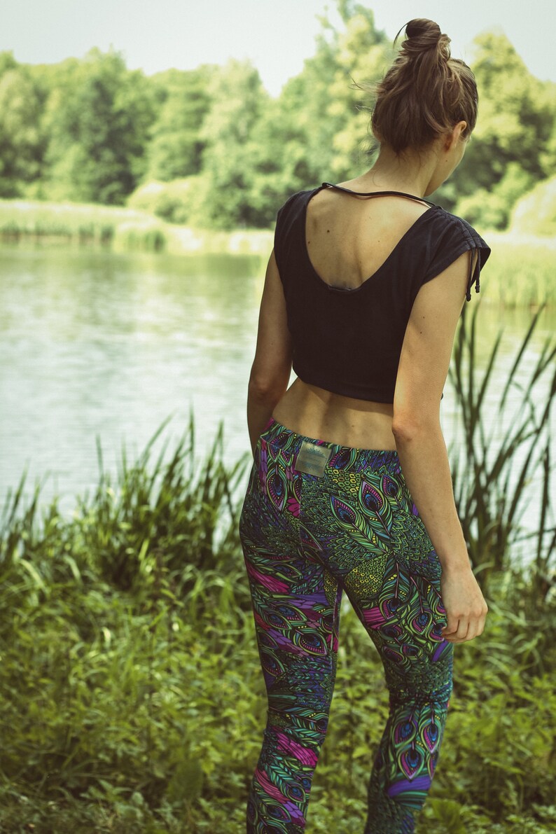 May include: A person wearing a black crop top and patterned leggings. The leggings feature a vibrant peacock feather design in shades of green, purple, and pink. The background shows a lake and lush greenery.