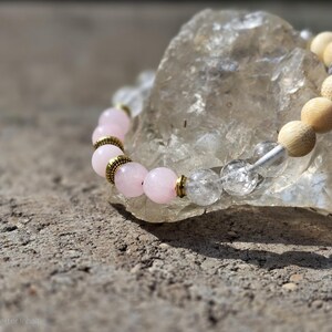 Könnte beinhalten: Ein Armband mit Perlen aus Rosenquarz, Bergkristall und Holzperlen. Das Armband hat goldfarbene Akzente und wird auf einem klaren, grob geschnittenen Stein präsentiert. Die Perlen haben einen Durchmesser von etwa 1 cm.