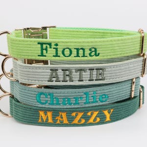 May include: Four corduroy dog collars in green, grey, and teal, stacked on top of each other. Each collar has a gold-coloured metal clasp and ring. The names "Fiona", "Artie", "Charlie", and "Mazzy" are embroidered on the collars in different colours.