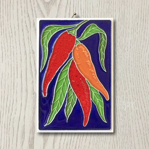 Könnte beinhalten: Eine blaue Keramikfliese mit weißem Rand zeigt ein Design aus drei roten und orangen Chilischoten mit grünen Blättern auf dunkelblauem Hintergrund.