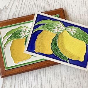 Può includere: Due sottopentola in ceramica a forma di limone su sfondo blu e verde. I limoni sono gialli con fiori bianchi e foglie verdi.