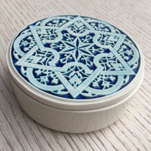 May include: A white ceramic trinket box with a blue and white geometric design on the lid. The design features a starburst pattern with intricate details.