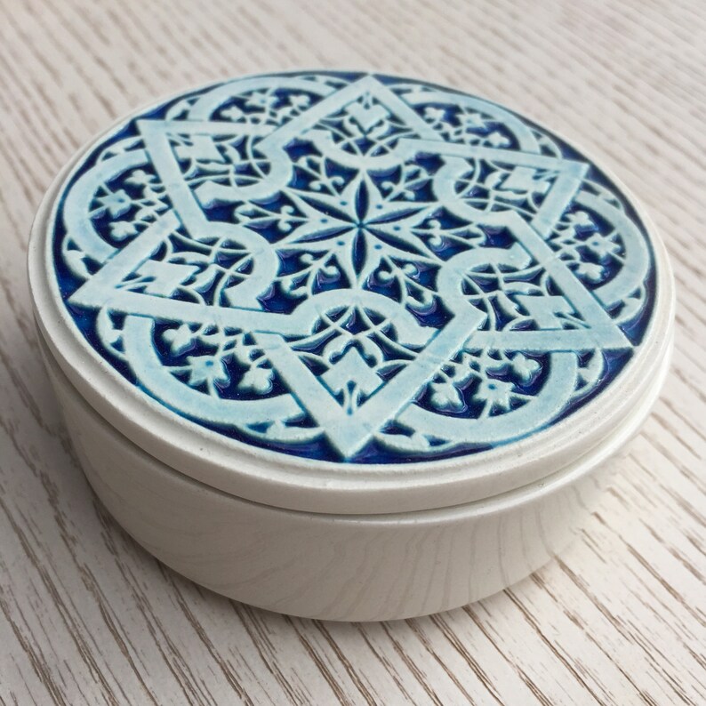 May include: A white ceramic trinket box with a blue and white geometric design on the lid. The design features a starburst pattern with intricate details.