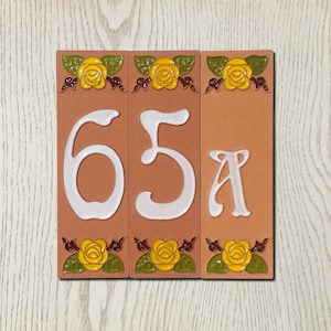 May include: Three terracotta tiles with white numbers 65A and yellow roses with green leaves. The tiles are arranged horizontally.