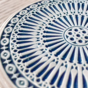 May include: A white ceramic coaster with a blue and white geometric design. The design features a central circle with a repeating pattern of eight circles, surrounded by a ring of radiating lines.