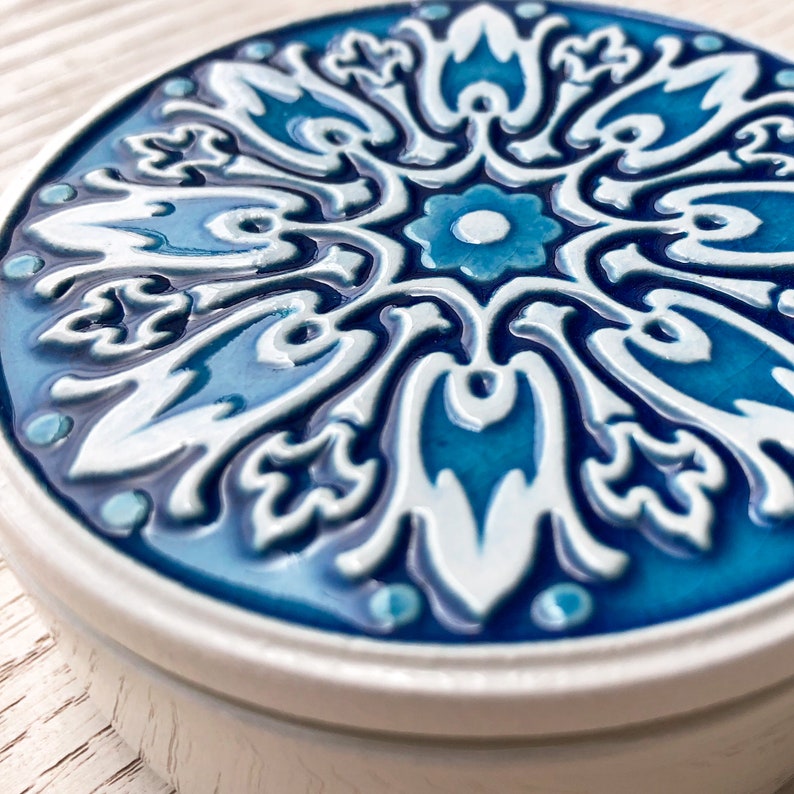May include: A round ceramic trinket box with a blue and white floral design. The box has a raised, white floral pattern on a blue background.