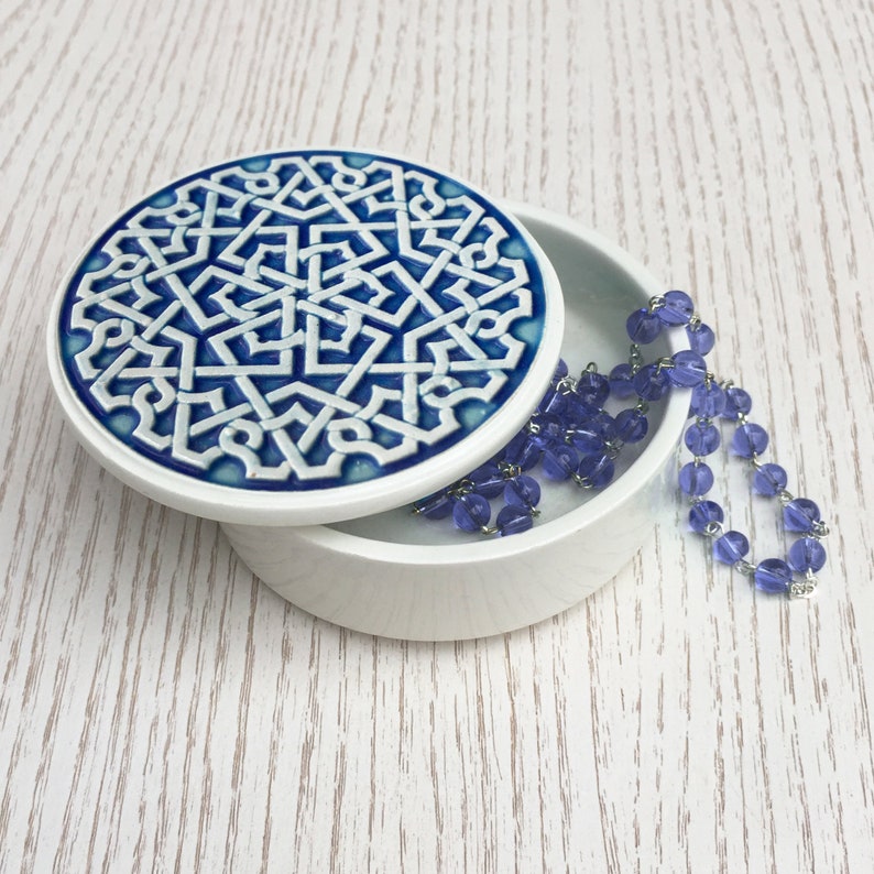 May include: A white ceramic trinket box with a blue and white geometric design on the lid. The box is open and a strand of purple beads is spilling out.