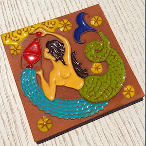 Peut inclure: Un carreau en terre cuite avec une illustration colorée d'une sirène versant de l'eau d'un pichet. Le carreau porte les mots "Acqua Rio" écrits en jaune dans le coin supérieur gauche.