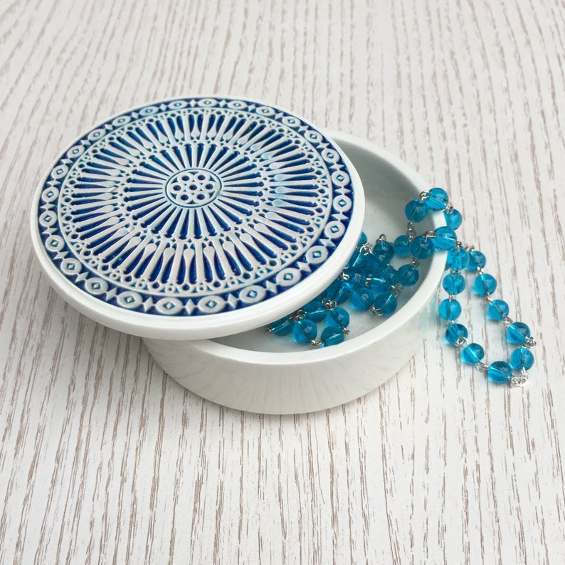 May include: A white ceramic trinket box with a blue and white geometric design on the lid. The box is open and a turquoise blue beaded necklace is inside.